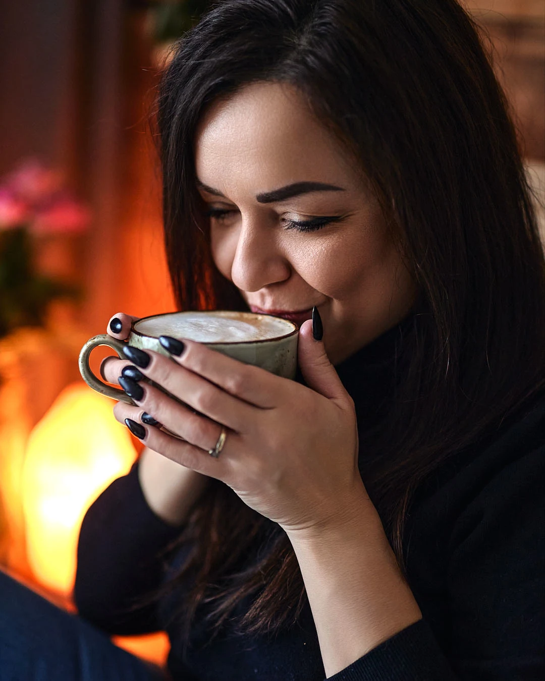 Sylwia Karolina Janiec, bilingual Polish teacher, holding a cappuccino beside a warm glowing lamp in an intimate café setting